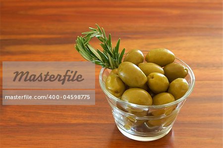 Single jar of green olives with stick of rosemary on wooden table background with copy space
