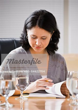 A young woman is seated at a desk in an office. She is looking down at a book.  Vertically framed shot.