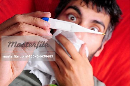 A sick man with a fever, checking his temperature with an electric thermometer. His mouth is covered by a tissue paper and his eyes are big and shocked at the temperature on the thermometer. The pillow he's laying on is red.