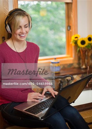 A young woman is working on a laptop in her kitchen.  She is wearing headphones, smiling, and looking away from the camera.  Vertical.