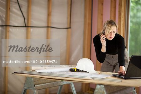 A young woman is looking at a laptop and talking on the phone.  She is working at a construction site.  Horizontally framed shot.