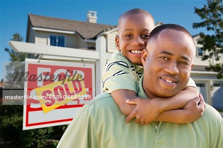 Happy African American Father and Son in Front of New Home and Real Estate Sign.