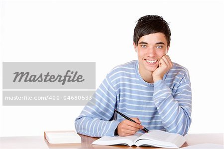 Young student is sitting on desk with open book and learns for his exams. He looks smiling into camera. Isolated on white.