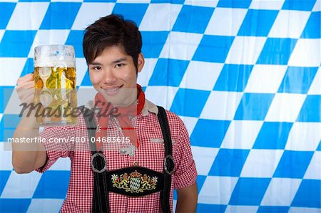 Asian Boy is holding a full Oktoberfest beer stein and smiles happy in camera. In background Bavarian flag visible.