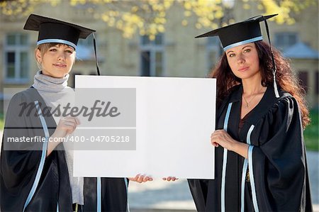 Two graduates hold a clean sheet in sunlight, with blurred college building in the background