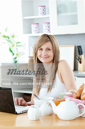 Smiling woman having breakfast in front of the laptop looking at the camera in the kitchen