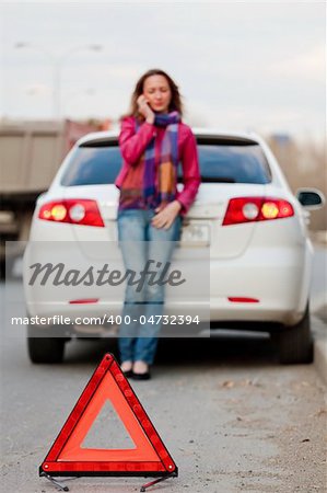 Woman calls to a service standing by a white car. Focus is on the red triangle sign. Evening light.