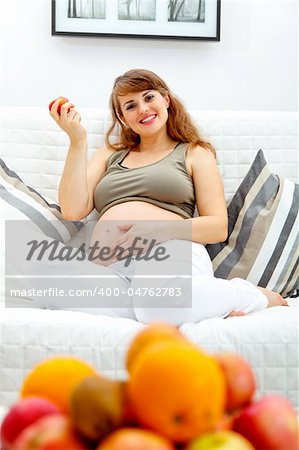 Smiling  beautiful pregnant woman sitting on divan at home and  holding fruit in hand