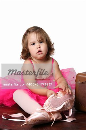 Little girl wearing a pink ballet outfit sitting next to an antique suitcase trying on oversized ballet shoes