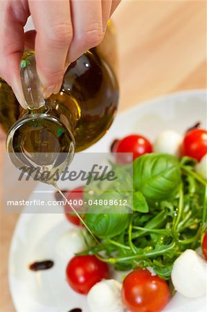 Pouring olive oil salad dressing onto tomato, mozarella and rocket salad with basil garnish and balsamic vinegar