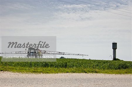 Rural landscape with the sky, a field, a water tower and a tractor spraying crops