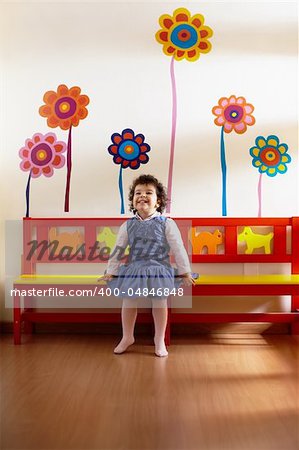 hispanic female preschooler sitting on bench in kindergarten and smiling. Vertical shape, full length, front view