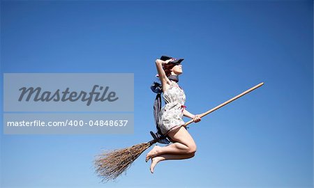 Young red-haired witch on broom flying over green grass field