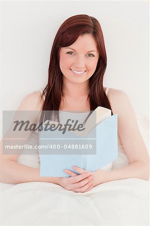 Good looking red-haired female reading a book while sitting on her bed