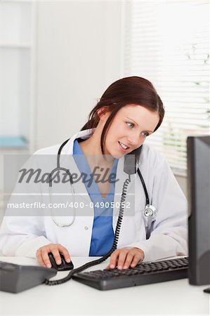 A female doctor is working with her pc in an office whilst telephoning