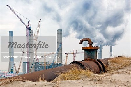An old oil pipe with valve and fosset being replaced by a new powerplant, being constructed in the background. Next to it, the old, fuming chimneys of the current powerplant