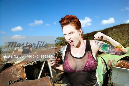 Angry tattooed Caucasian woman yelling with fists clenched in junkyard.