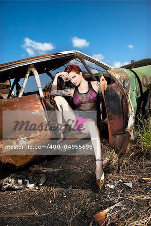 Sexy tattooed Caucasian woman sitting seductively in old rusted car in junkyard.