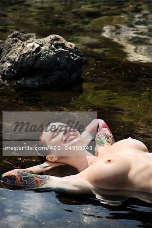 Sexy nude tattooed Caucasian woman lying and smiling in tidal pool in Maui, Hawaii, USA.