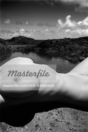 Close up torso of young adult nude Caucasian woman lying on boulder on rocky Maui coast.