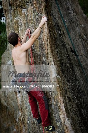 A male climber, viewed from above, climbs a very high and steep crag.  The image is taken as the climber clips into the bolt.