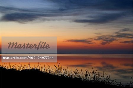 Early morning. The sun soon will rise above the sea and will illuminate clouds. In the quiet sea beautiful reflection. Pleasant colours. A quiet nature. Silhouettes of a grass on a coast