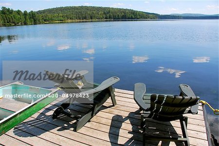 Two adirondack wooden chairs on dock facing a blue lake with clouds reflections