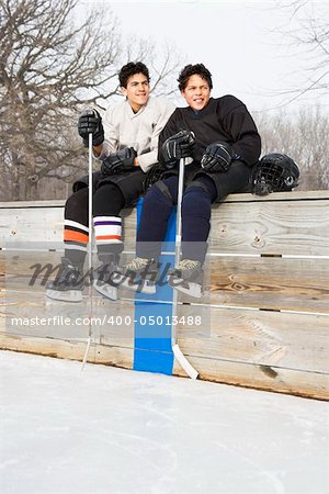 Two boys in ice hockey uniforms sitting on ice rink sidelines looking and smiling.