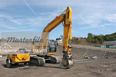 Industrial yellow digger standing idle on a building site with a small red diesel storage vehicle to the side. Set against a blue sky.