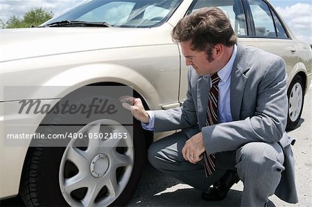 A businessman on the road with a flate tire.  He has just discovered the screw that caused the tire to go flat.