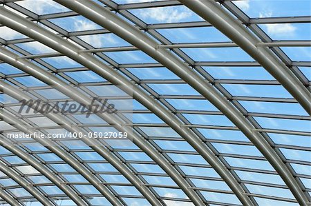 Silver metal curved roof joists in a conservatory with glass paned windows in between and a blue sky and clouds beyond.