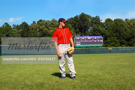 young teen male stands on baseball field in red navy uniform.  He is looking sideways.  Scoreboard is in background of picture.