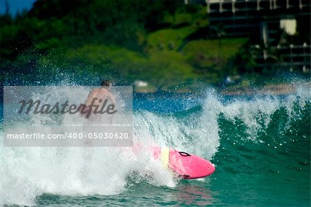Surfer is surrounded by splashing and crashing wave and wipes out.  His board is pink and he has on red swim trunks.