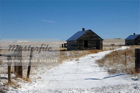 Deserted homestead with barbed wire across driveway describes this rural home in Colorado.  Empty windows and open doorway.  Blue sky and snowy ground.