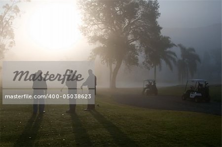 Early morning golfers silhouetted in a dense fog with a rising sun