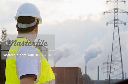 A man dressed in hardhat and safety hi-visibilty jacket surveys a factory with chimneys putting out smoke