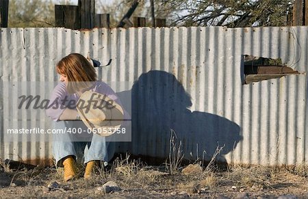 Woman in a Cowboy hat sitting against a Corrugated Metal fence and Looking to her Right.