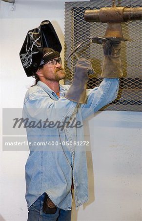 Welder hammering a piece of metal he's been welding.  Focus on the man's face.  (helmet was customized by the welder - the designs are not trademarked or copyrighted)