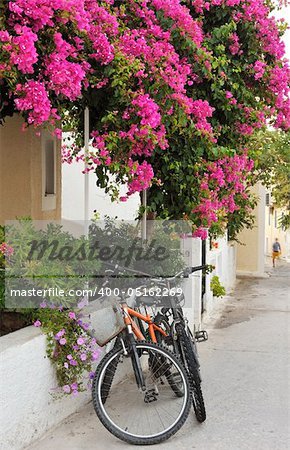 Scene from a narrow alley from a Greek island village, showing two bicycles leaning against a house covered in colorful flowers