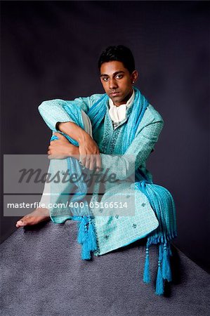 Beautiful authentic Indian hindu man in typical ethnic groom attire sitting relaxed on top of rock leaning on knee. Bangali male wearing a light blue agua decorated Dhoti with shawl.