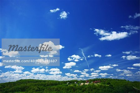 Image of a blue sky with the clouds on a sunny day and some rural houses in the green forest.