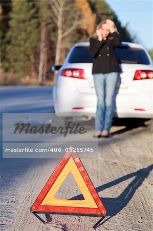 Young woman standing by her damaged car and calling for help. Focus is on the red triangle sign.