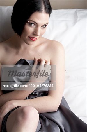 Sexy naked young caucasian adult woman with red lips, short black hair and a pierced eyebrow, covered in a dark satin sheet and sitting on a bed