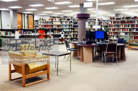 Computer station at the university college library with seating area in the foreground. One young female student searching far in the background.