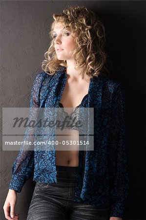 sexy blond curly haired woman in blue shirt and bra against a black wall