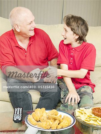 Father and son playfully fighting over a chicken wing as they watch the football game.