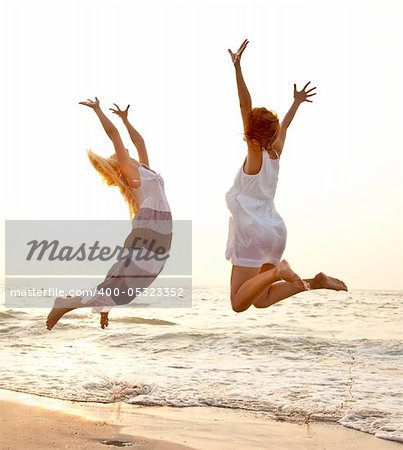 Two beautiful young girlfriends jumping on the beach at sunset. Photo with counter-light on background.