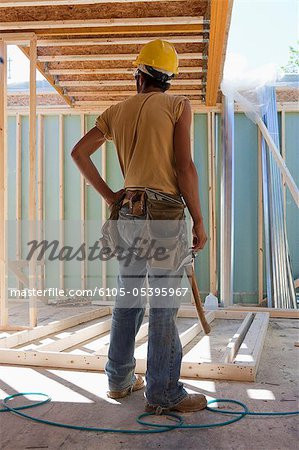 Hispanic carpenter with a hammer standing next to wall frame at a house under construction