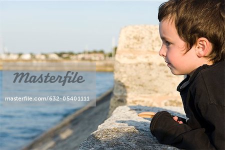 Little boy leaning against stone wall, looking at ocean view