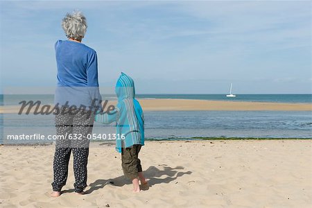 Grandmother and grandson standing together on beach, looking at boat on horizon